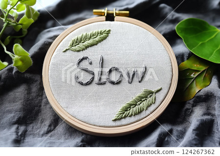 A wooden hoop adorned with embroidery art. It features the word "Slow" crafted in gray thread with surrounding green leaf stems, evoking a sense of mindfulness and calm. Slow living, hand made concept 124267362