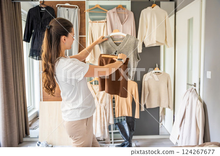 Young woman holds and compares clothing in front of a wardrobe filled with hanging outfits in a bedroom. Choosing to wear is part of personal style, daily fashion decisions, and wardrobe organization 124267677