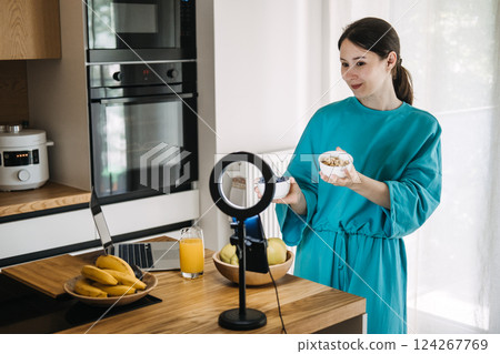 Young Asian woman filming healthy food content with smartphone and ring light in kitchen. Creator economy and wellness influencer trends 124267769