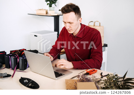 Young man managing handmade jewelry brand on laptop in modern craft workspace. Trends: personal branding, maker identity, digital entrepreneurship Young man managing handmade jewelry brand on laptop in modern craft workspace. Trends: personal branding, maker identity, digital entrepreneurship 124267806
