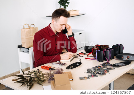 Young White male entrepreneur on phone call while assembling handmade jewelry products at home workspace. Trends: remote entrepreneurship, craft-based microbusiness, creative gig economy Young White male entrepreneur on phone call while assembling handmade jewelry products at home workspace. Trends: remote entrepreneurship, craft-based microbusiness, creative gig economy 124267807