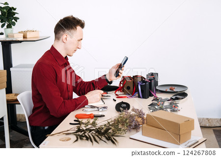 Young Eastern European man photographing handmade jewelry packaging with smartphone at craft workspace. Trends: product photography, craft e-commerce, digital selling Young Eastern European man photographing handmade jewelry packaging with smartphone at craft workspace. Trends: product photography, craft e-commerce, digital selling 124267808