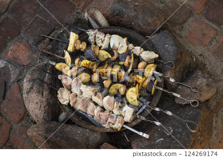 Skewers of marinated meat and vegetables are grilling over an open flame, creating a smoky aroma in a rustic outdoor environment with a stone surface. Skewers of marinated meat and vegetables are grilling over an open flame, creating a smoky aroma in a rustic outdoor environment with a stone surface. 124269174