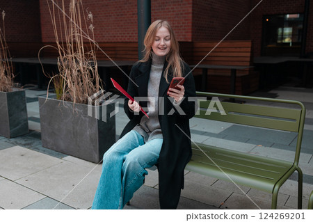 Office worker with a red paper folder sits on a bench and looks at his phone, social media, streaming and news, Internet, technology and digital with business female in outdoors for communication Office worker with a red paper folder sits on a bench and looks at his phone, social media, streaming and news, Internet, technology and digital with business female in outdoors for communication 124269201