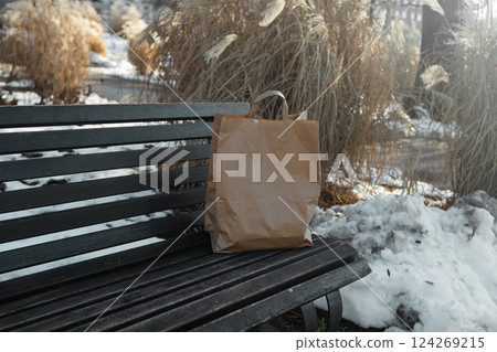 A paper craft bag with Takeaway food products groceries stands on a wooden bench in a winter sunny day. Fast delivery 124269215