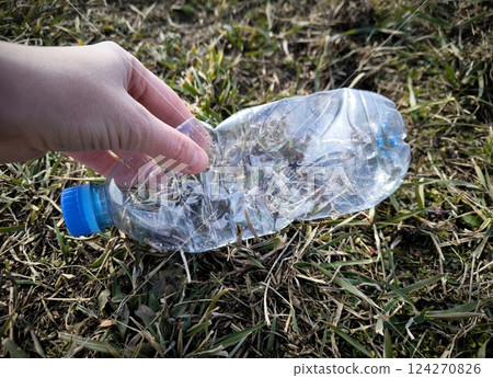 crumpled, twisted plastic bottle lies in grass, environmental pollution, close-up. plastic bottle on the grass is garbage. hand picks up trash from the grass in the park. crumpled, twisted plastic bottle lies in grass, environmental pollution, close-up. plastic bottle on the grass is garbage. hand picks up trash from the grass in the park. 124270826