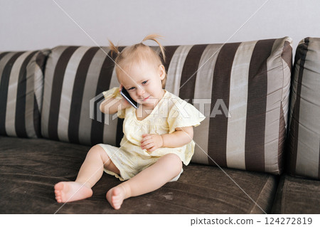 Cheerful infant girl holding smartphone to ear, mimicking phone call. Joyful child in light-colored outfit sitting on couch showcasing curiosity and imagination talking using smartphone at home. 124272819
