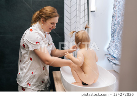Caring mother gently cleansing cheerful toddler daughter in contemporary bathroom sink, sharing intimate bonding moment during bathtime routine. Concept of parenting love and care. 124272826