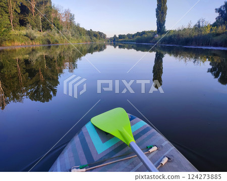 SUP board with green paddle on surface of river water with trees on banks on calm windless summer evening. Person swimming on SUP board on pond with reflective water surface. Tourism travel rest sport SUP board with green paddle on surface of river water with trees on banks on calm windless summer evening. Person swimming on SUP board on pond with reflective water surface. Tourism travel rest sport 124273885
