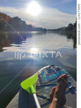 Bare legs of woman on SUP board on surface of river or lake with sun reflecting and with trees on banks on warm summer evening. Person swimming on SUP board on pond on calm day. Tourism rest sport 124273891