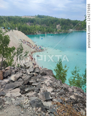 Many people swimming in the blue water near the sandy shore in a flooded quarry on a summer day. Beautiful blue water of large lake and many people swimming near the sandy shore of the slope. Top view 124273898