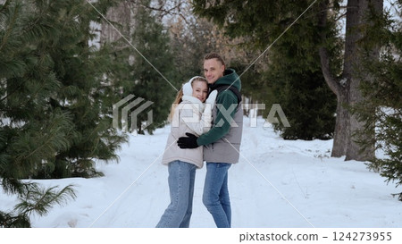 Smiling couple embracing while walking through snow covered evergreen forest, expressing love and connection amid winter landscape's serene natural beauty 124273955