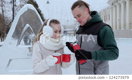 Winter clad couple sharing hot drinks, standing close to intricately carved ice sculpture, expressing joy and intimacy during cold seasonal moment 124273979