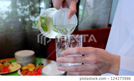 Uniformed server pouring chilled lemonade into crystal glassware, surrounded by fresh fruits and empty plates inside elegant dining establishment 124273986