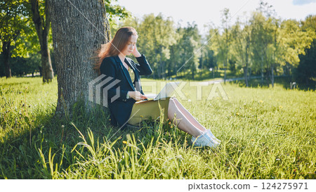 Young businesswoman using laptop while sitting under tree in park, enjoying fresh air and nature during work break Young businesswoman using laptop while sitting under tree in park, enjoying fresh air and nature during work break 124275971