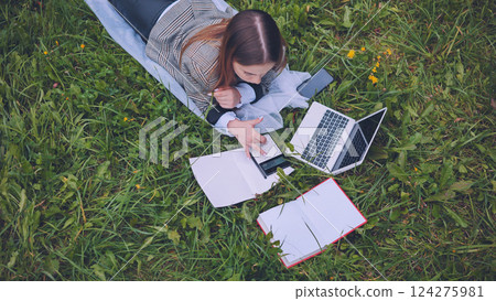 Student resting on verdant lawn, working on math assignment with laptop, basking in sunlight and natural surroundings 124275981