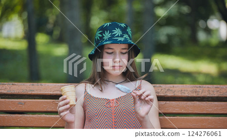 Young female with cannabis themed bucket hat savoring frozen treat at sunny urban green space 124276061