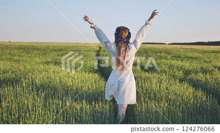 Young woman wearing flower wreath walking in green wheat field with arms outstretched enjoying freedom and nature 124276066