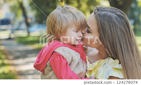 Young mother kissing her toddler daughter on the cheek in a park, expressing love, care, and happiness 124276074