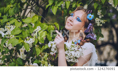 Young woman with creative makeup and butterfly in hair posing near blooming lilac tree in spring garden 124276091