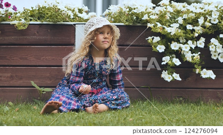 Young girl sitting barefoot on garden grass, wearing sunhat, clutching delicate white blossom 124276094