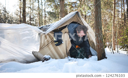 Homeless man crawling out of makeshift tent in a snowy forest during a frigid winter day, facing the harsh realities of survival Homeless man crawling out of makeshift tent in a snowy forest during a frigid winter day, facing the harsh realities of survival 124276103
