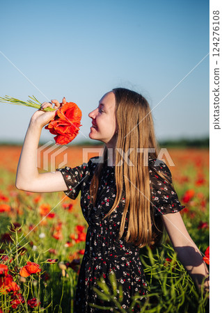 Woman inhaling fragrance of red poppies, long brown hair flowing, standing in golden sunset lit wildflower meadow Woman inhaling fragrance of red poppies, long brown hair flowing, standing in golden sunset lit wildflower meadow 124276108