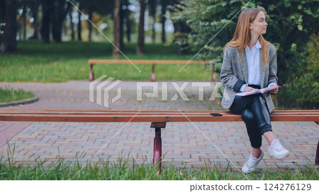 Young woman reading book, relaxing on urban park bench, surrounded by greenery and soft sunlight 124276129