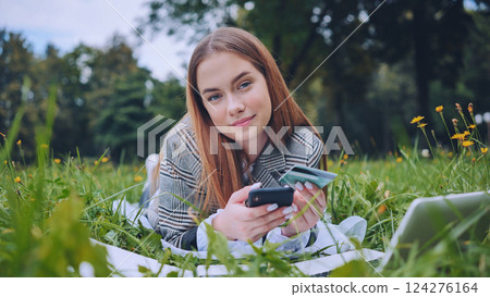 Woman relaxing on green lawn, browsing online store with mobile device and payment card 124276164