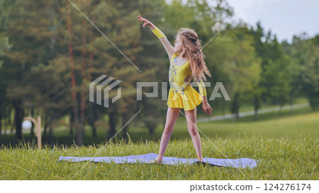 Rhythmic gymnast practicing flexible pose on yoga mat, extending arm gracefully in outdoor green park setting Rhythmic gymnast practicing flexible pose on yoga mat, extending arm gracefully in outdoor green park setting 124276174