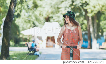 Young woman riding electric scooter, smiling freely while cruising through sunlit park, wearing trendy summer attire Young woman riding electric scooter, smiling freely while cruising through sunlit park, wearing trendy summer attire 124276179