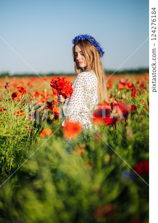 Blonde female posing with cornflower crown, carrying poppy bouquet amid vivid flowering meadow Blonde female posing with cornflower crown, carrying poppy bouquet amid vivid flowering meadow 124276184
