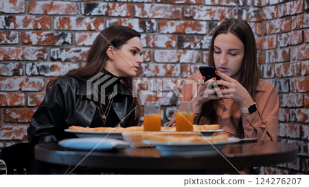 A girl peeks into her friend's smartphone at a coffee shop. A girl peeks into her friend's smartphone at a coffee shop. 124276207