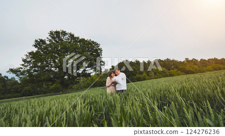 Loving parents holding newborn amid golden wheat, peaceful oak tree backdrop creating serene family landscape at sunset 124276236