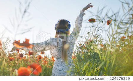 Young woman wearing flower crown, polka dot dress, standing amid blooming poppies with arms spread wide, embracing scenic landscape and natural beauty 124276239
