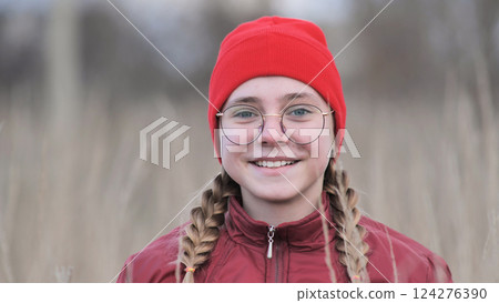 Teenage girl with braided hair, red beanie, glasses, smiling while standing amid tall grass on sunny day Teenage girl with braided hair, red beanie, glasses, smiling while standing amid tall grass on sunny day 124276390