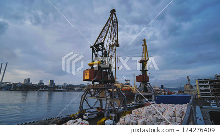 Cargo cranes handling goods in a commercial port at sunset, with cloudy sky and cityscape in background Cargo cranes handling goods in a commercial port at sunset, with cloudy sky and cityscape in background 124276409