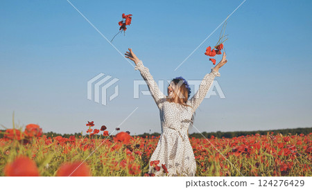 Young woman wearing white dress and flower crown throws poppy flowers in a field, enjoying summer 124276429