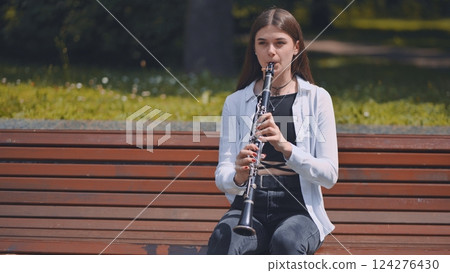 Young woman performing clarinet while sitting on park bench, surrounded by verdant landscape with trees and foliage Young woman performing clarinet while sitting on park bench, surrounded by verdant landscape with trees and foliage 124276430