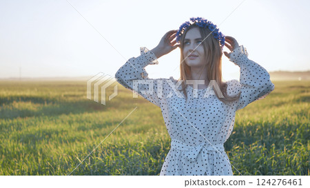 Young woman with flower wreath adjusting her hair while standing in a field at sunset, enjoying the peace and beauty of nature 124276461