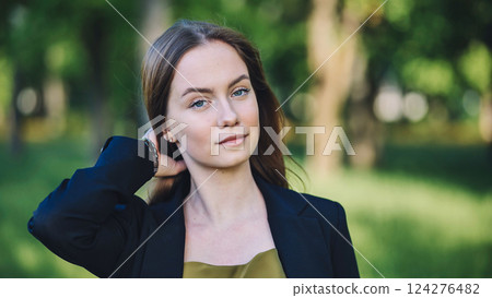 Professional businesswoman smoothing hair, standing in green park, relaxing during midday work break, sunlight highlighting confident posture 124276482