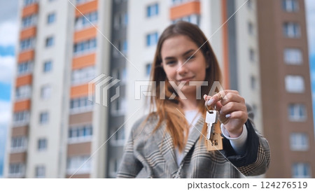 Female professional holding house keys, standing near modern apartment building, representing property ownership and real estate investment success Female professional holding house keys, standing near modern apartment building, representing property ownership and real estate investment success 124276519