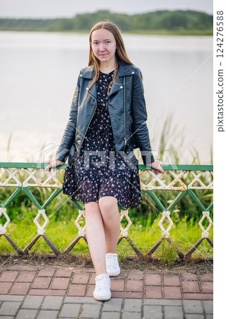 Teen girl wearing a floral dress and black leather jacket sitting on a rusty fence by the lake enjoying the view Teen girl wearing a floral dress and black leather jacket sitting on a rusty fence by the lake enjoying the view 124276588
