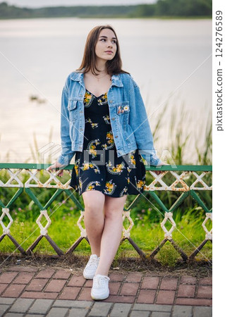 Young woman leaning on lakeside fence, wearing floral dress and denim jacket, basking in golden sunset light Young woman leaning on lakeside fence, wearing floral dress and denim jacket, basking in golden sunset light 124276589