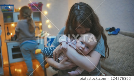 Loving mother tenderly holding newborn, gently kissing infant's head while sibling plays nearby in cozy home setting Loving mother tenderly holding newborn, gently kissing infant's head while sibling plays nearby in cozy home setting 124276593