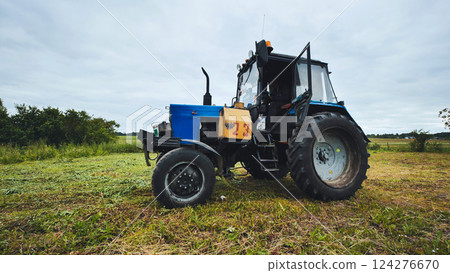 Blue tractor staying parked in a mowed field under cloudy sky 124276670