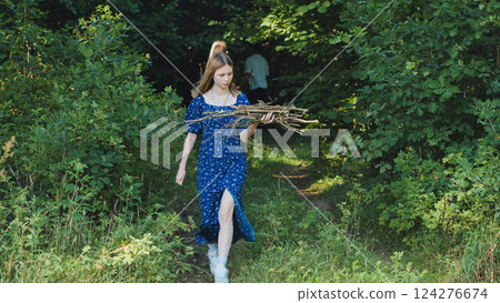 Blonde girl in blue dress carrying firewood in forest, enjoying camping adventure with friends 124276674