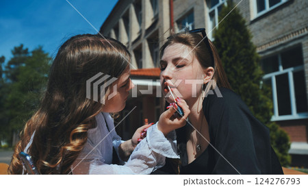 Two friends bonding and sharing beauty tips while applying lip gloss outside on a sunny day during a school break Two friends bonding and sharing beauty tips while applying lip gloss outside on a sunny day during a school break 124276793