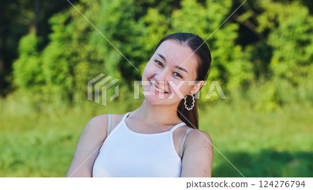 Happy teen girl with long brown hair in a white top and earrings smiling in a summer park Happy teen girl with long brown hair in a white top and earrings smiling in a summer park 124276794