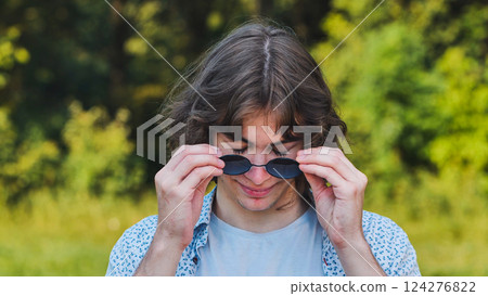 Cheerful young man playfully lowering his sunglasses and smiling in a sunny park setting Cheerful young man playfully lowering his sunglasses and smiling in a sunny park setting 124276822